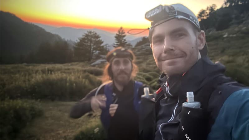 Two trail runners wearing headlamps and hydration packs take a selfie at sunrise or sunset in a mountainous landscape, with grassy terrain and trees in the background.odo