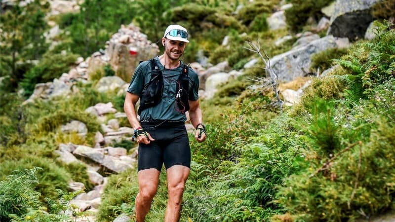 Trail runner wearing a cap and hydration vest smiling while running along a rocky, green mountain path.