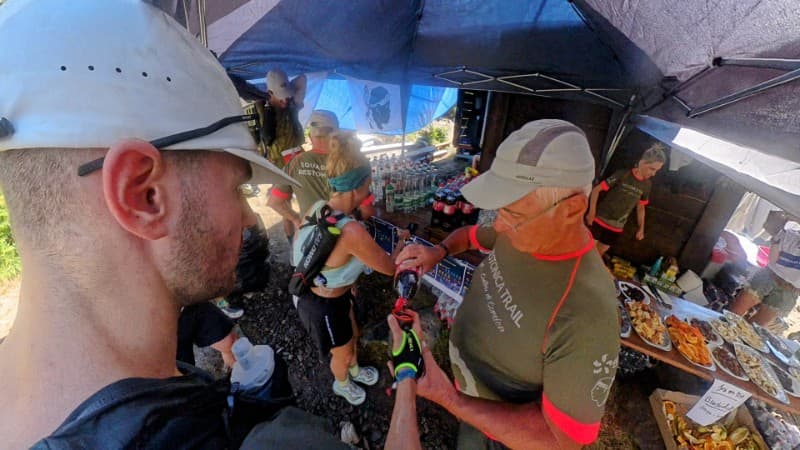 A trail runner smiles while jogging through a lush, green mountain path surrounded by rocks, ferns, and dense vegetation, wearing a cap, hydration vest, and sportswear.