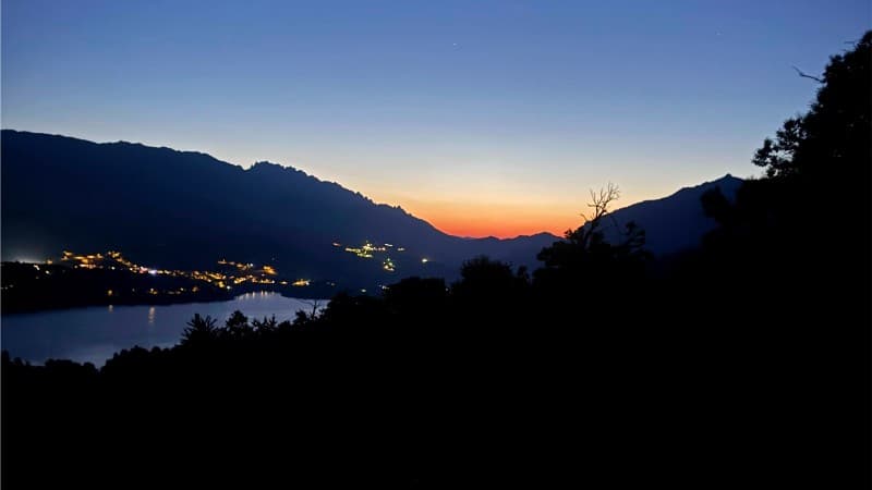 A peaceful mountain landscape at dawn or dusk, with a lake reflecting the last light of the sky, village lights scattered along the shore, and mountains silhouetted against a blue and orange horizon.