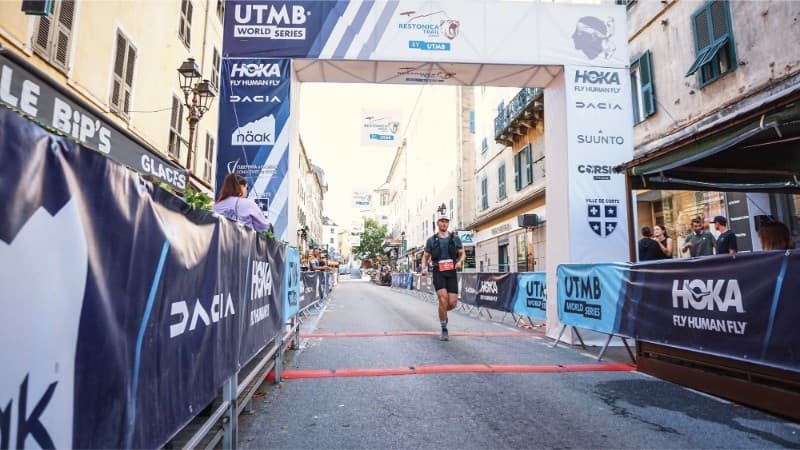 A trail runner crosses the finish line at a UTMB World Series race in a city street, with banners, spectators, and event branding visible on the barriers and archway.