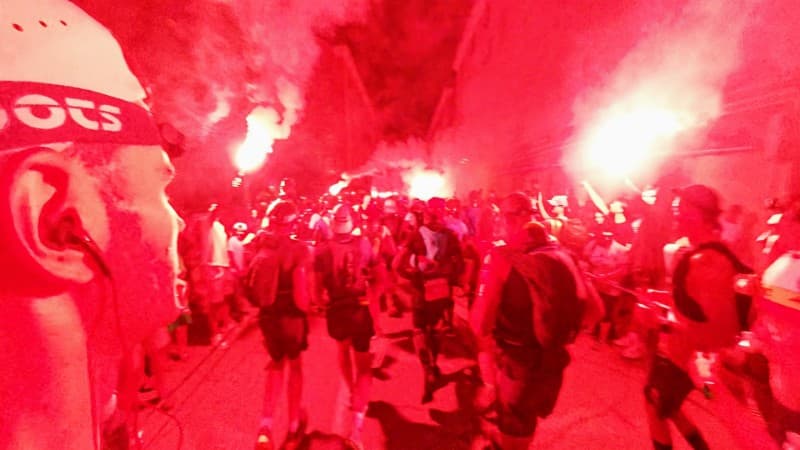 A group of runners at night, surrounded by a crowd holding bright red flares, with smoke filling the street and the scene illuminated in red light, creating an intense and energetic atmosphere at the start of a race or event.