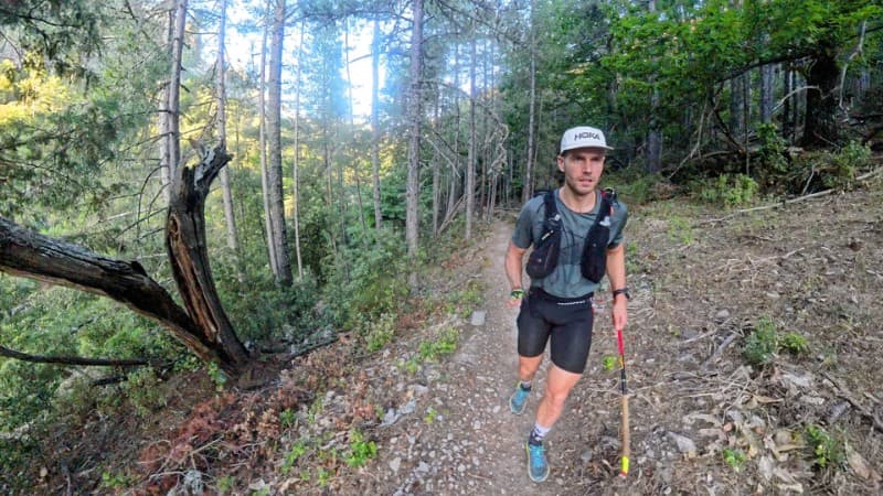 Un traileur portant une casquette et un gilet d'hydratation trottine sur un sentier étroit et rocailleux à travers une forêt dense aux grands arbres et à la végétation abondante.