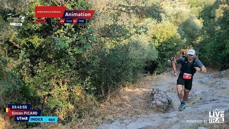 Trail runner wearing a white cap and race bib running uphill on a rocky path surrounded by dense green vegetation.