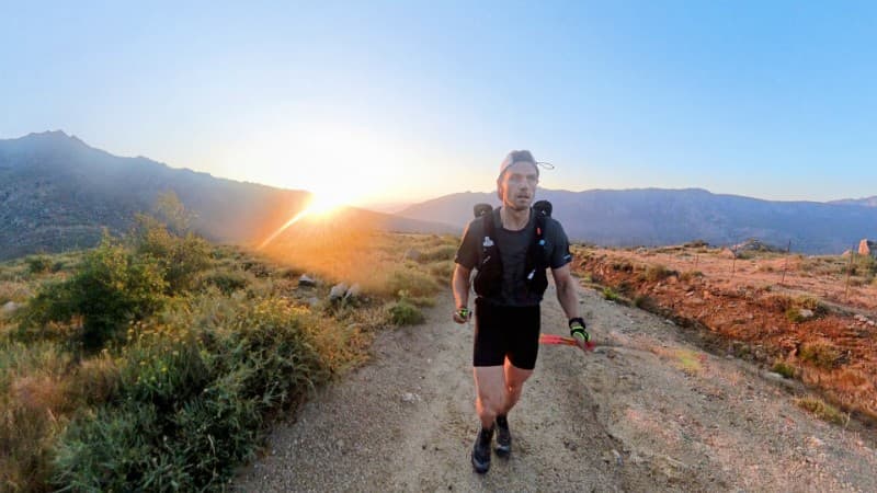 A trail runner wearing a hydration vest and athletic gear runs on a dirt path through a mountainous landscape at sunrise, with the sun rising over distant hills and casting a warm glow across the scene.