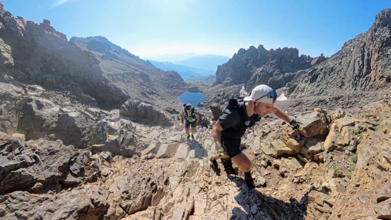 Un traileur gravit un terrain rocheux et escarpé en montagne sous un ciel bleu limpide, suivi d'un autre coureur, avec un petit lac alpin visible au loin en contrebas.