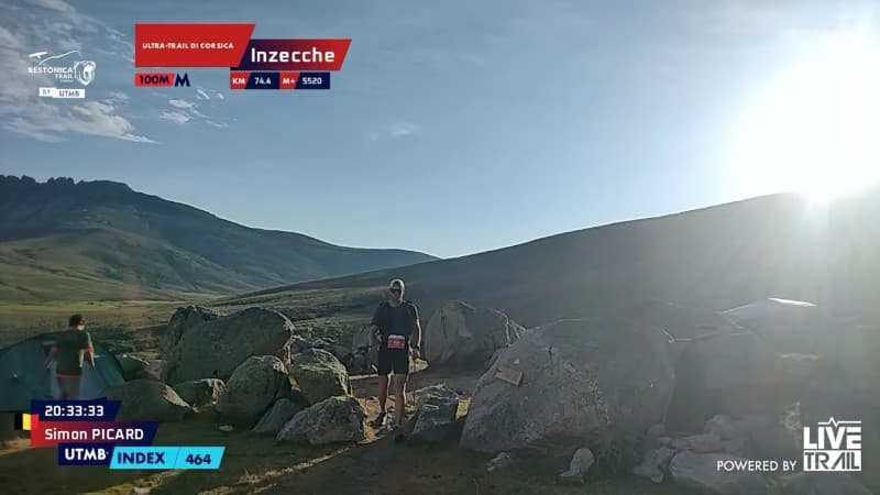 Trail runner standing among large rocks on a mountain path at sunrise with hills in the background.