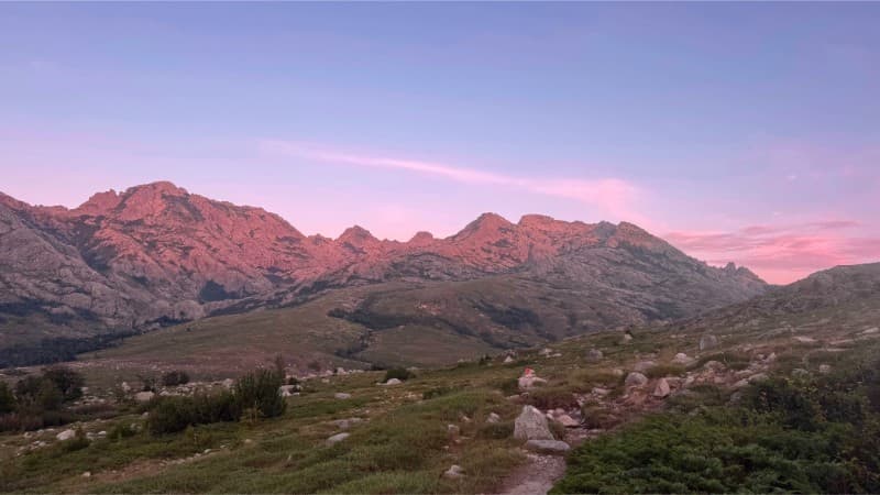 Un paysage de montagne pittoresque au crépuscule, avec des sommets escarpés baignés d'une douce lumière rose sous un ciel dégagé, des prairies herbeuses et un terrain rocheux au premier plan.