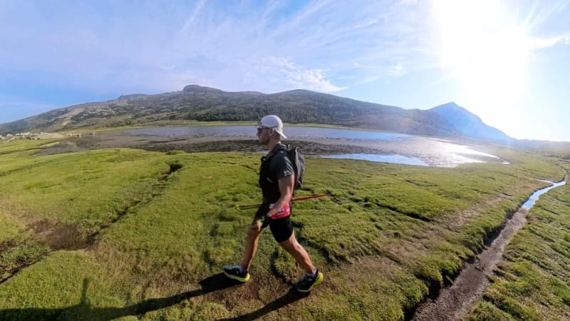 A trail runner wearing a cap and hydration vest walks across a grassy, open plain near a reflective lake, with mountains in the background and bright sunlight in the sky.