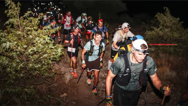 A group of trail runners wearing headlamps and backpacks hike up a rugged, rocky path at night, surrounded by bushes and illuminated by beams of light in the darkness.