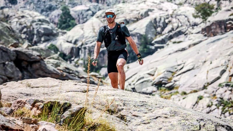 A trail runner wearing sunglasses, a cap, and a hydration vest walks over rocky terrain in the mountains, with rugged cliff faces and boulders in the background.