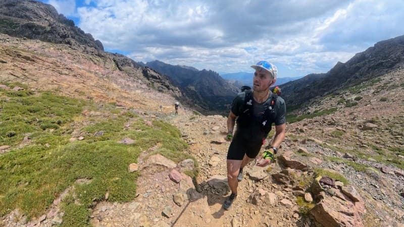 A trail runner wearing a cap and hydration vest climbs a rocky mountain path under a partly cloudy sky, with rugged peaks and green patches of grass in the background.